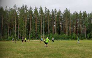 Jalkapallo-ottelu käynnissä, pelaajia metsän laidalla sijaitsevalla nurmikentällä.