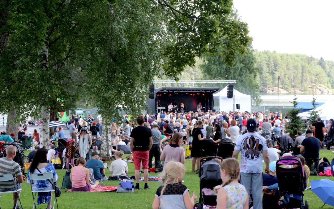 Satoja eri ikäisiä ihmisiä kesäisessä puistossa kuuntelemassa musiikkia, joka tulee taustalla olevalta lavalta.
