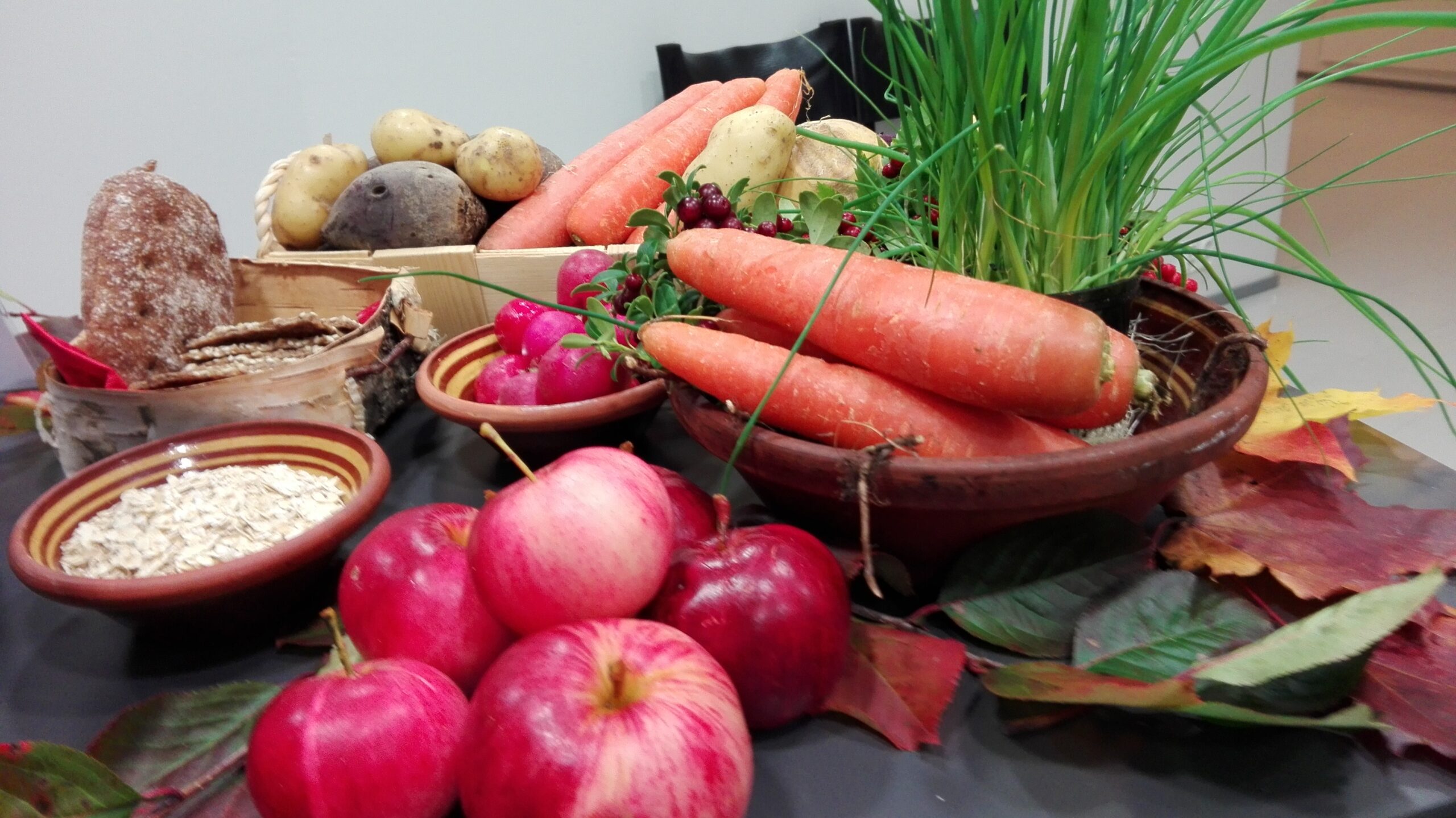 Porkkanoita, omenia, puolukoita, retiisejä ja muita kasviksia sekä leipää ja kaurahiutaleita pöydällä ja kulhoissa.