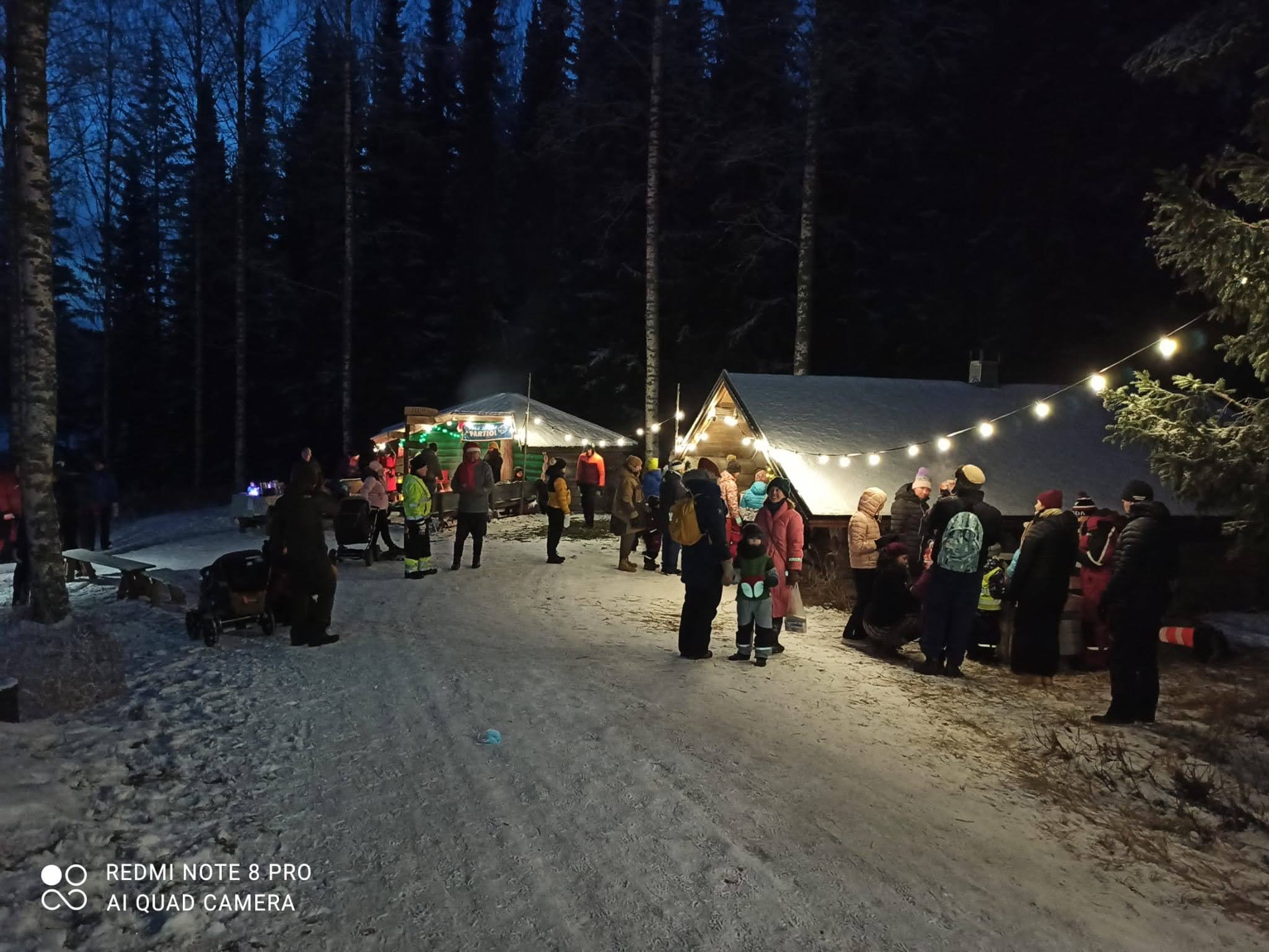 Lumisen tien varrella jouluvaloja, paljon ihmisiä, aitta ja laavu.