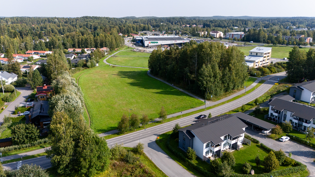 Ilmakuva tyhjästä tontista, jonka ympärillä teitä ja puita, taustalla kauppakeskus.