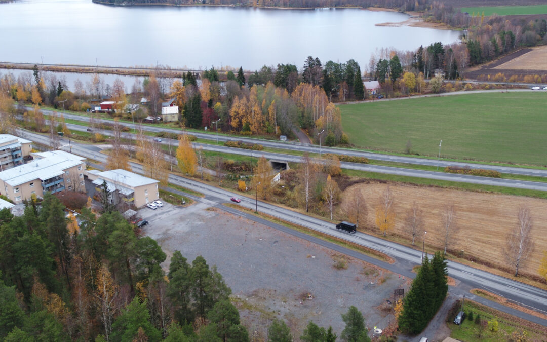 Ilmakuva tyhjästä tontista, jonka ympärillä on teitä, taloja ja peltoa, taustalla järvi.